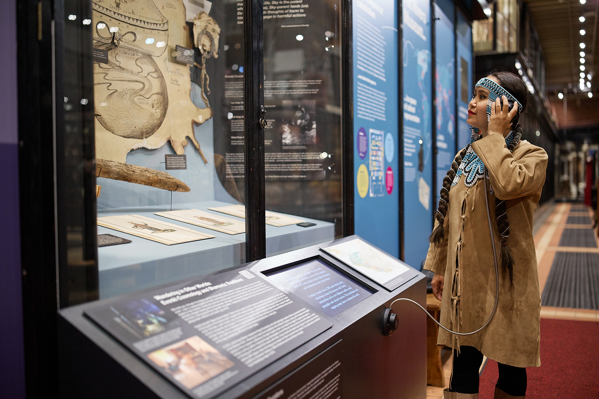 A woman stands smiling while looking at a display case showing objects from Evenkia in Sibera, holding a listening device to her ear.