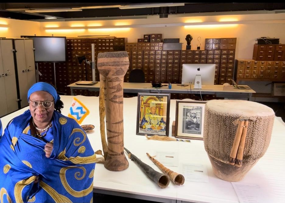 A smiling lady dressed in a blue and gold wrapper stands next to a tall thin drum, two horns and a short stout drum. In the background are photographs of two men.