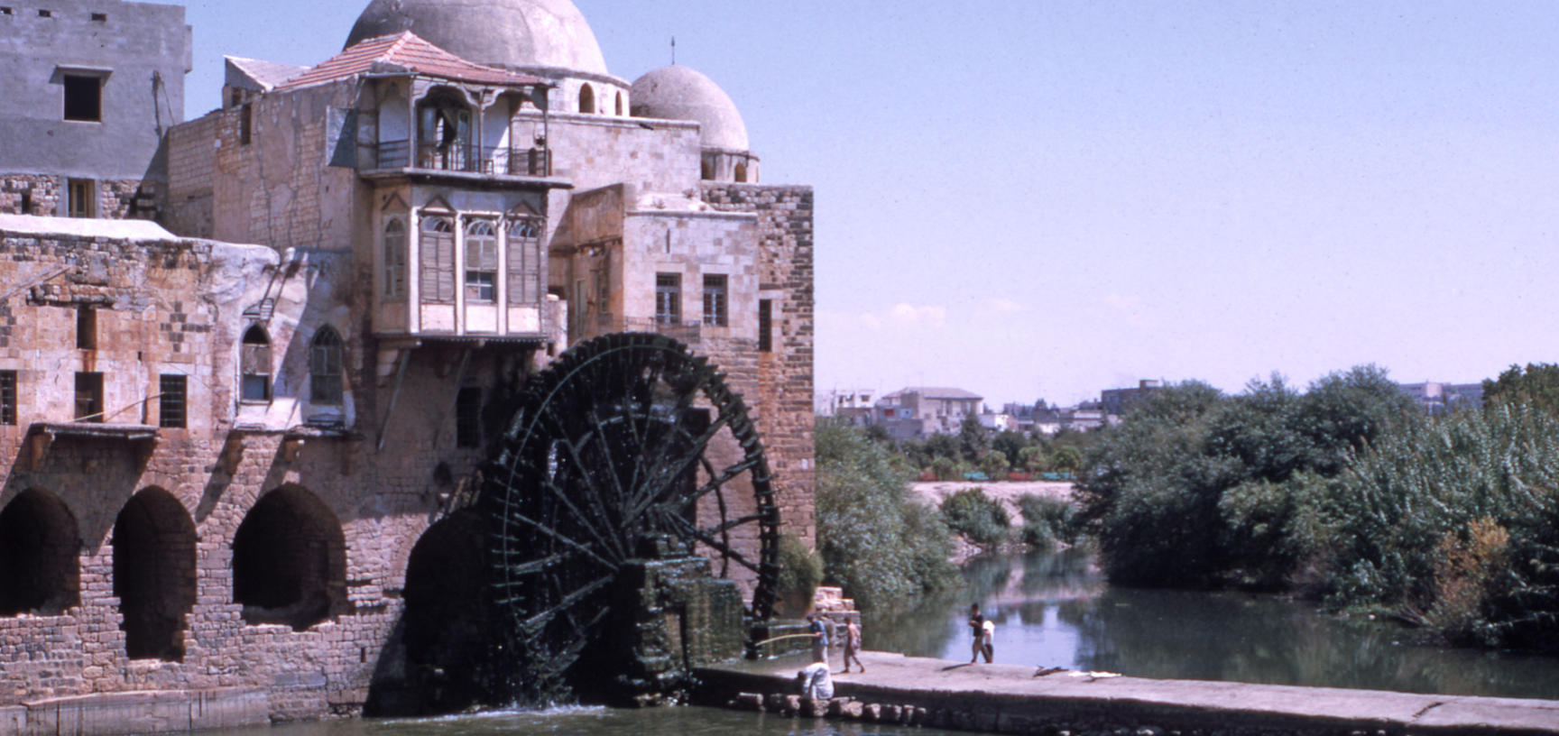 A waterwheel attached to a building rotates over a river.  