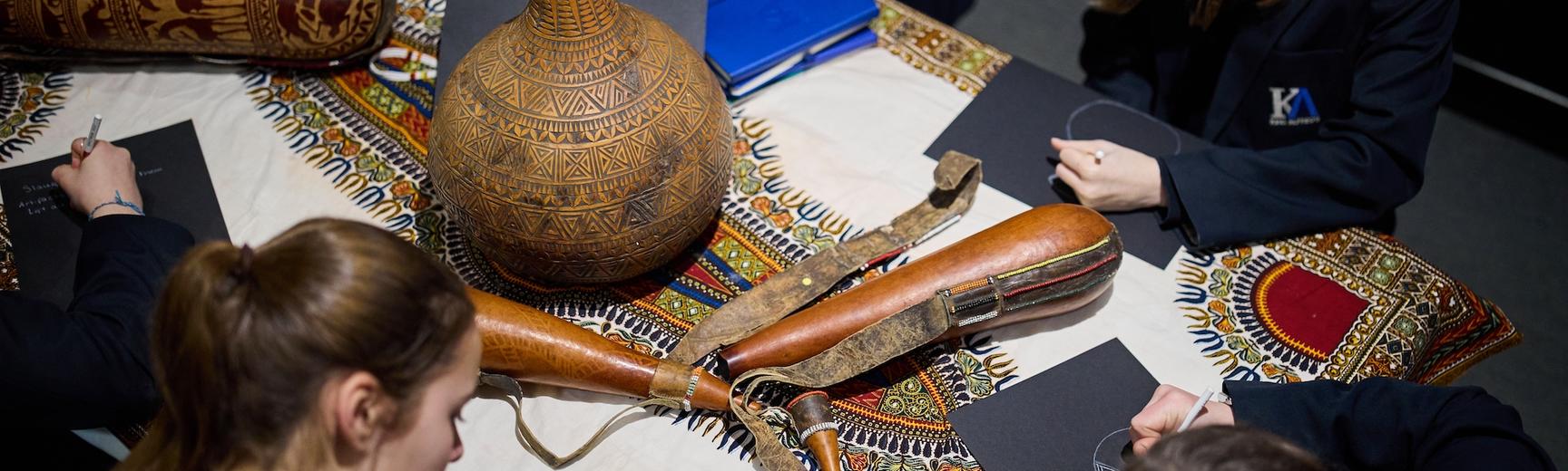 School students sitting around table drawing decorative gourds and textiles which are grouped on the table.