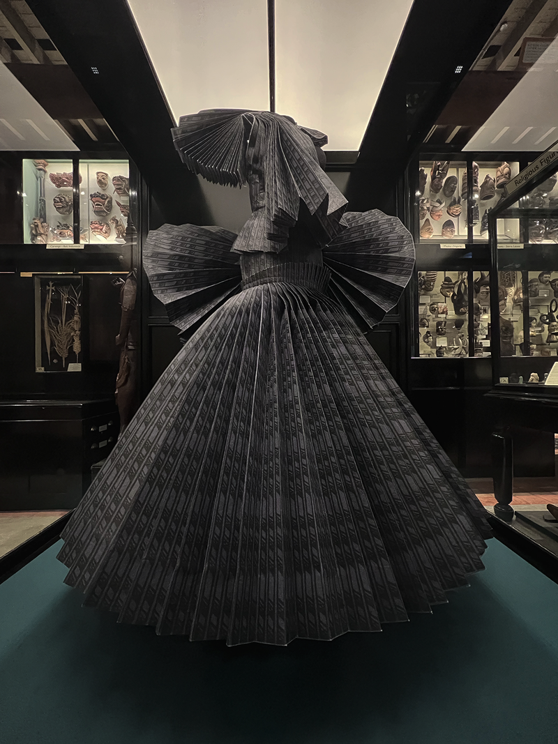 Sculptural dress made from pleated, printed fabric on display in a glass case surrounded by museum exhibits 