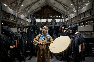Woman wearing blue beaded headdress beating drum in front of people dressed in crow costumes