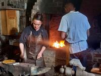 Woman hammering iron on an anvil in front of a man tending a fire in a brick waist-height furnace.